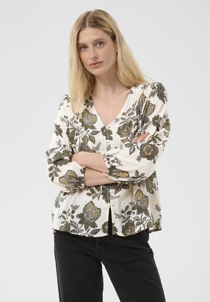 Femme blonde, bras croisés, portant un chemisier blanc à fleurs boutonné et un pantalon noir sur un fond uni.