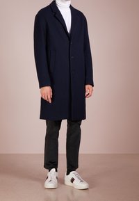 Marineblauer Wollmischung-Mantel mit Reverskragen, Drei-Knopf-Verschluss, seitlichen Taschen, kombiniert mit schwarzen Hosen und weißen Sneakers mit bunten Akzenten.