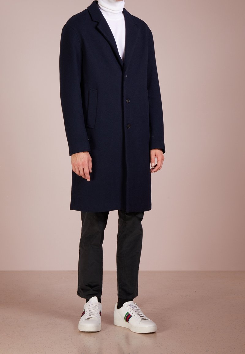 Marineblauer Wollmischung-Mantel mit Reverskragen, Drei-Knopf-Verschluss, seitlichen Taschen, kombiniert mit schwarzen Hosen und weißen Sneakers mit bunten Akzenten.