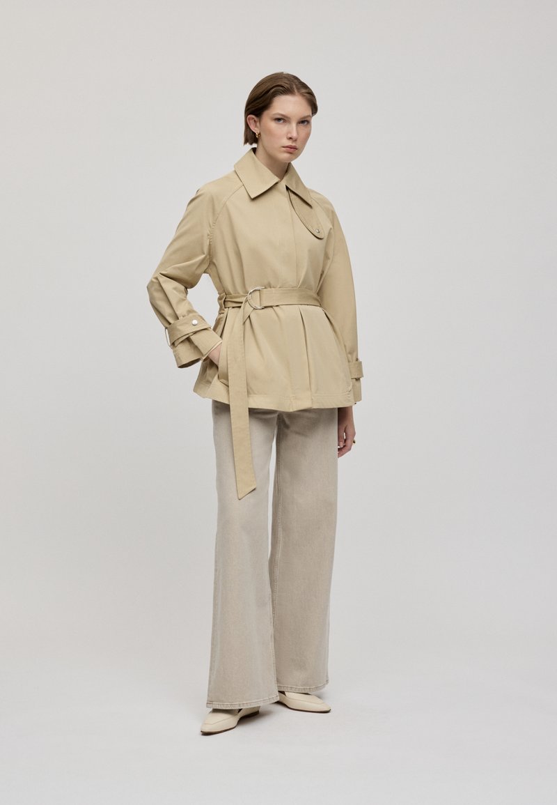 Beige Trenchcoat mit Gürtel, Kragen und Schulterklappen, kombiniert mit hellbeigen, weit geschnittenen Hosen und cremefarbenen Schuhen.