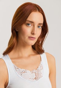 Weißes Tanktop mit einem floralen Spitzenausschnitt in aufwendiger Stickerei. Der Stoff wirkt glatt mit einer sanften Textur und verstellbaren Trägern.