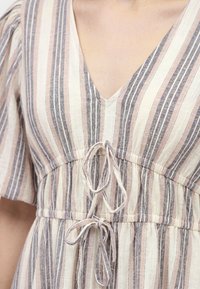 Gros plan d'un chemisier rayé avec des lignes verticales beige, grises et roses, avec un col en V et deux nœuds noués au niveau de la poitrine et de la taille.