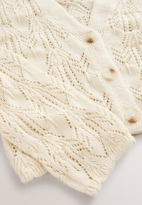 Cardigan en maille couleur crème avec motifs losanges et dentelle, doté de trois boutons beiges sur la fermeture frontale.