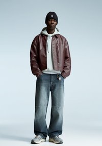 Veste en cuir bordeaux sur un sweat à capuche gris, associée à un jean bleu à jambes larges. Bonnet en tricot noir. Baskets avec des accents gris et beige.
