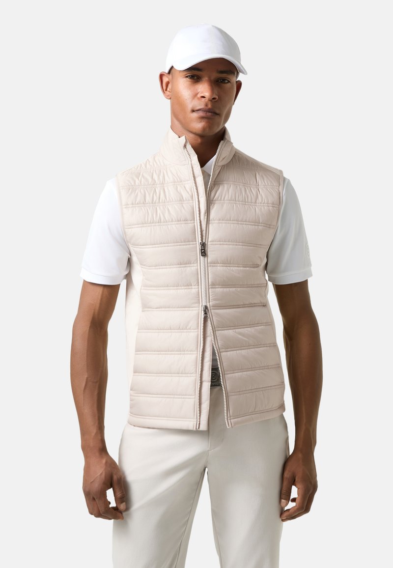 Helle beige gepolsterte Weste mit horizontalen Nähten, Frontreißverschluss und hohem Kragen, die über einem weißen Poloshirt mit kurzen Ärmeln getragen wird.