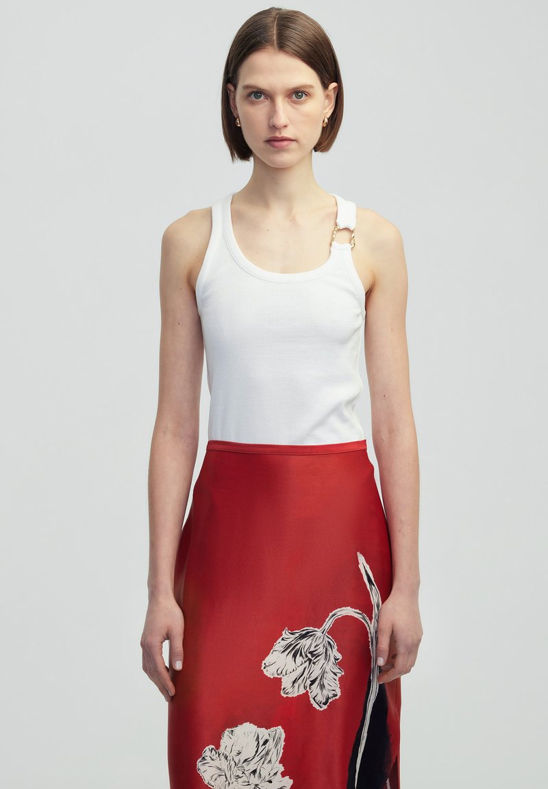 Witte geribde tanktop met verstelbare gouden hardware detail op één schouderband, gecombineerd met een rode rok met zwart-witte bloemenpatronen.