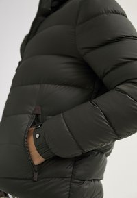 Dunkelgrüne Steppjacke mit gestepptem Design, seitlichen Reißverschlusstaschen und glatter Textur. Merkmale sind elastische Bündchen und ein Stehkragen.