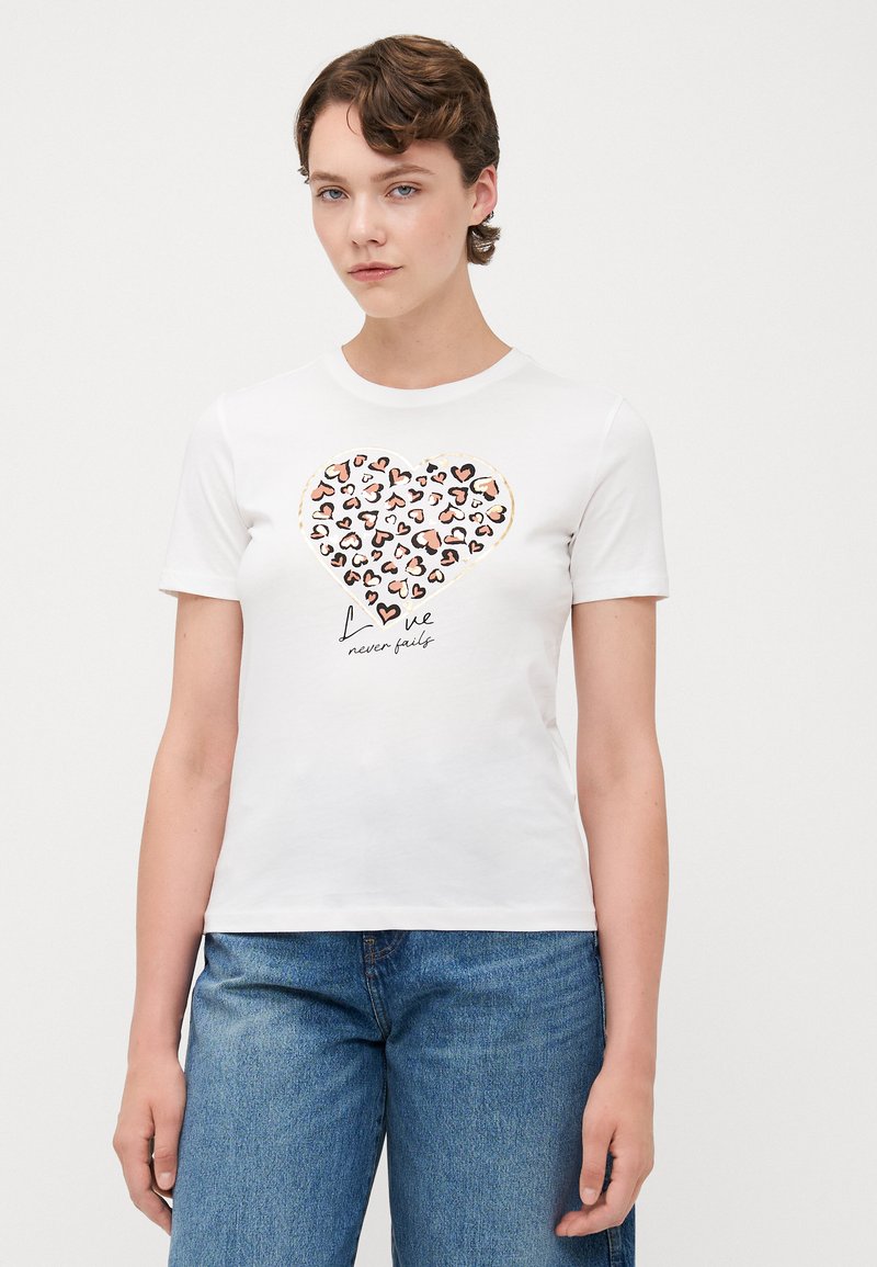 Hvid t-shirt med korte ærmer med hjerteprint, der viser forskellige farvede hjerter og teksten "Kærlighed fejler aldrig" nedenfor. Fremstillet af bomuld.