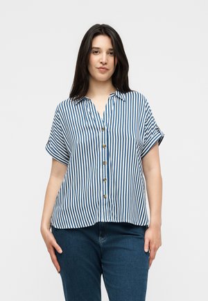 Kvinne iført kortermet blå og hvit vertikalstripet skjorte med knapper og mørkeblå jeans, stående foran en ensfarget lys bakgrunn.