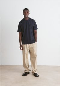 Polo shirt navy cu mânecă scurtă, asociat cu pantaloni largi bej și pantofi slip-on negri; expus pe un fundal neutru.