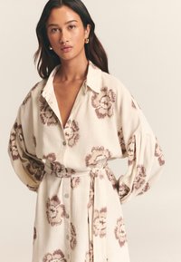 Balta lininė suknelė su rudais gėlių raštais, mygtukų užsegimu priekyje ir juosmens diržu. Ilgos rankovės ir per didelis sukirpimas.