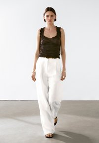 Brown ruched tank top with scalloped edges paired with white wide-leg trousers. Model wears flat black sandals and simple metal bracelets.