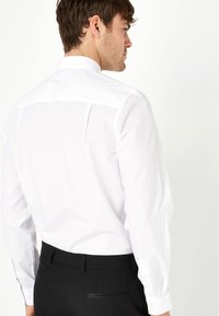 Weißes Langarmshirt mit Stehkragen, das eine glatte Textur und knöpfbare Manschetten aufweist, kombiniert mit schwarzen Hosen.