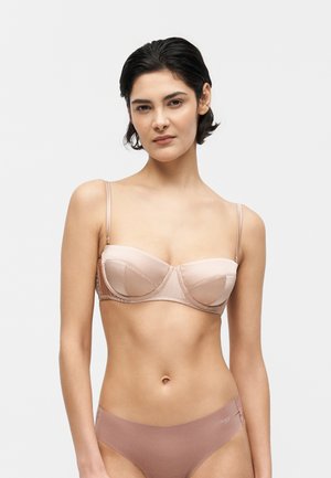 Femme aux cheveux courts et foncés portant un soutien-gorge rose clair sans bretelles et un slip taille haute assorti sur un fond clair uni.