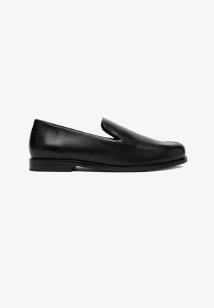 Mocasín negro de cuero tipo slip-on con tacón bajo y punta redondeada, mostrado de perfil sobre un fondo blanco.