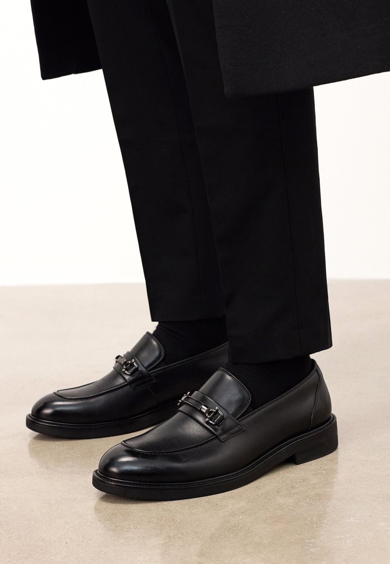 Black men's loafers made of smooth leather, featuring a decorative strap with metallic accents, paired with black trousers and socks.