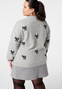 Femme aux cheveux noirs attachés en queue de cheval, portant un pull gris avec des motifs de nœuds noirs et une jupe en chevrons grise, debout devant un fond blanc.