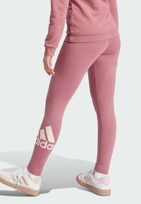 Leggings deportivos rosas con una textura suave, que presentan un gran logo blanco de Adidas en el tobillo. Combinados con zapatillas blancas y rosas.