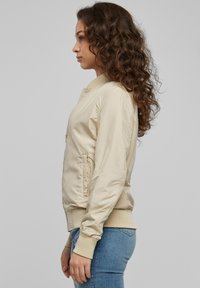 Bézs bomber dzseki sima, fényes felülettel, elöl cipzárral, oldalzsebekkel, valamint bordázott mandzsettákkal és szegéllyel. Kék farmernadrág fölött viselve.