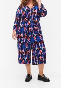 Jumpsuit con stampa animale multicolore in blu, rosa, rosso e nero. Maniche lunghe, vita con cintura, vestibilità ampia, pantaloni cropped e scarpe platform nere.