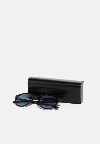 Lunettes de soleil noires avec des verres ronds, teinte bleue et accents en métal, positionnées à côté d'un étui noir brillant. Texture lisse.