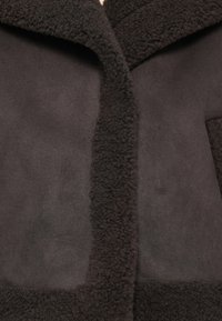 Läheltä kuvattu tumma ruskea shearling-takki, jossa on pehmeät mokkapaneelit ja paksu, karkea villareunus kauluksessa ja etuaukossa.