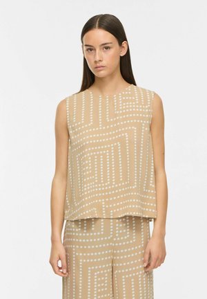 Femme portant un top beige sans manches et un pantalon assorti avec des motifs géométriques blancs en pointillés, debout devant un fond blanc uni.