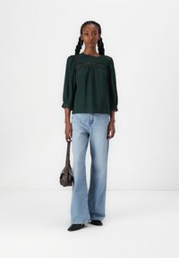 Blusa verde testurizzata con bordo in pizzo e maniche a tre quarti, abbinata a jeans svasati azzurri e una borsa a tracolla marrone.