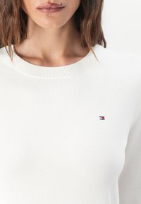Hvit strikket genser med rund hals; har et lite brodert logo i marineblått, rødt og hvitt på venstre bryst.