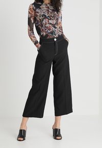 Mustat leveälahkeiset housut korkealla vyötäröllä, joissa on etutaskut ja valkoiset tikkaukset. Malli yhdistää ne kukkakuosisen pitkähihaisen paidan kanssa.