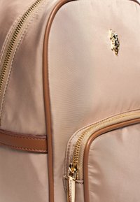 Sac à dos en nylon rose pâle avec des accents en cuir brun, doté de fermetures éclair dorées et d'un petit logo. Texture lisse avec une forme structurée et des poches.