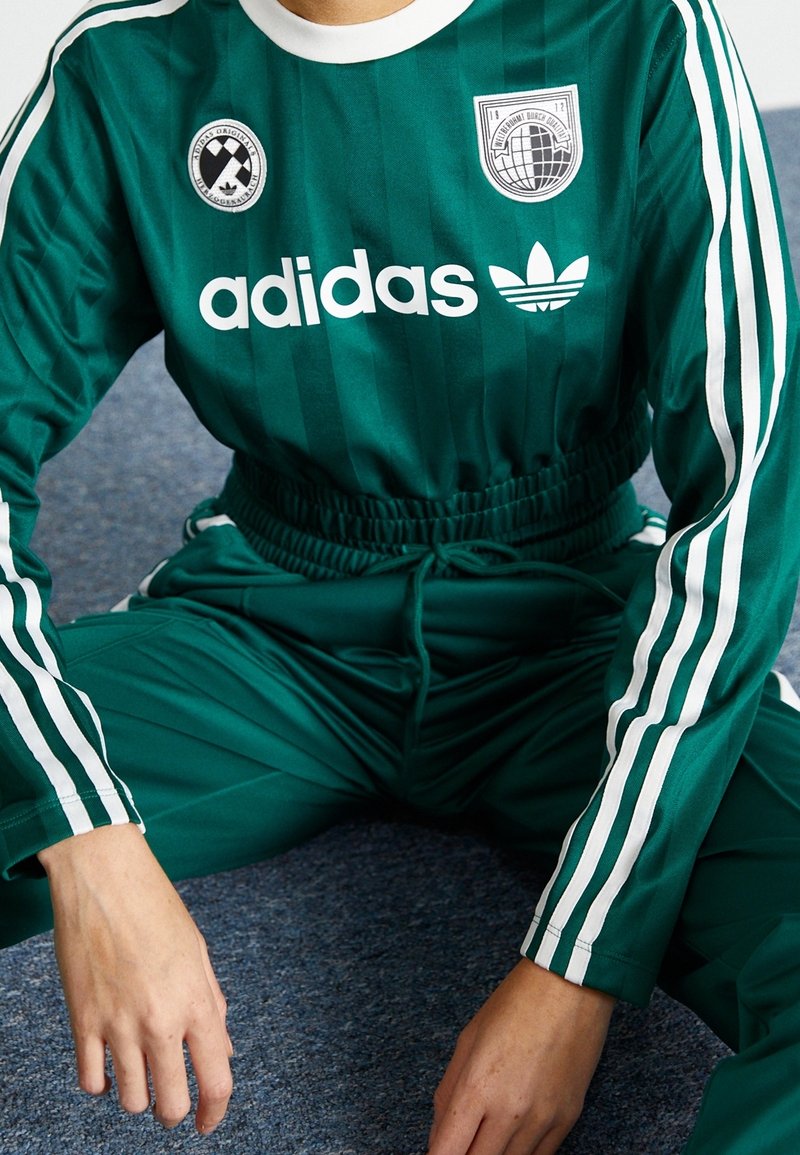 Grön Adidas träningsoverall med vita ränder, med elastisk midja och tydliga logotyper på bröstet. Slät, lättviktsmaterial.