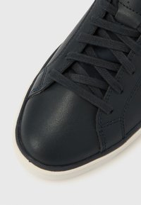 Κοντινό πλάνο μαύρου δερμάτινου αθλητικού παπουτσιού με μαύρα κορδόνια και λευκή λαστιχένια σόλα σε ανοιχτό φόντο.