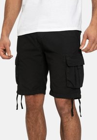 Mann, der schwarze Cargo-Shorts mit großen Seitentaschen und Kordelzügen trägt, kombiniert mit einem weißen T-Shirt, vor einem schlichten Hintergrund.