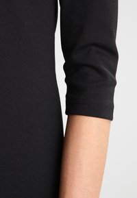 Bras portant une manche noire ajustée avec une couture visible sur un fond uni.