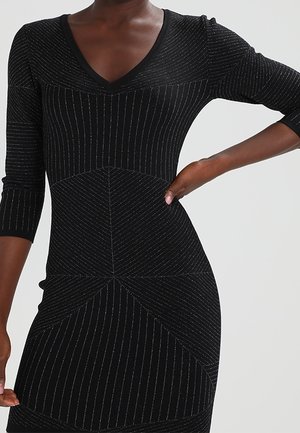 Kvinde iført en sort tætsiddende kjole med sølvfarvede, lodrette og diagonale nålestriber og trekvartlange ærmer, hånden hvilende på hoften.