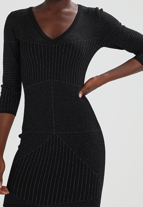 Femme portant une robe noire ajustée avec des fines rayures argentées verticales et diagonales, manches trois-quarts, main posée sur la hanche.