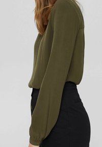 Blusa verde oliva con maniche lunghe e ampie e una vestibilità rilassata, abbinata a una gonna nera. Tessuto morbido e materiale leggero.