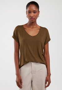 Oliwkowa, krótka koszulka z dekoltem w kształcie litery V oraz podwiniętymi rękawami. Wykonana z miękkiego materiału o luźnym kroju, zestawiona z jasnobeżowymi spodniami.