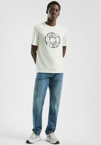 Jonge man staat met handen achter zijn rug, draagt een wit T-shirt met de tekst "Quite Sense", blauwe jeans en witte sneakers op een effen achtergrond.