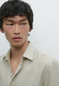 Collared shirt in light beige fabric, featuring a textured finish and a relaxed fit. Close-up view highlights buttons and collar details.