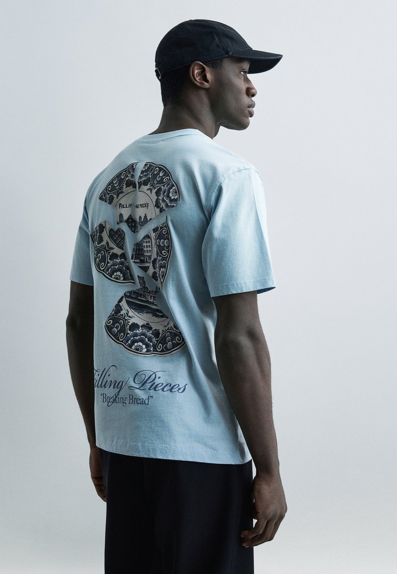 Άνδρας που φοράει ανοιχτό μπλε μπλ ουζάκι με γραφικό από ραγισμένο κεραμικό πιάτο και κείμενο «Filling Pieces Breaking Bread», μαύρο καπέλο και σκούρο παντελόνι.