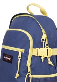 Zaino in nylon navy con accenti gialli, dotato di doppie cerniere, chiusura a fibbia e tasche laterali in rete. Logo Eastpak ben visibile.
