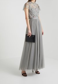 Graues Tüll-Maxikleid mit floraler Stickerei, durchsichtigen Puffärmeln und einem taillierten Oberteil. Kombiniert mit einer strukturierten schwarzen Clutch und sandaletten mit Riemchen.