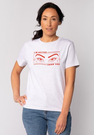 Femme aux cheveux foncés portant un T-shirt blanc avec des yeux stylisés rouges et le texte "JE SUIS MEILLEURE QUE TOI", associé à un jean bleu.