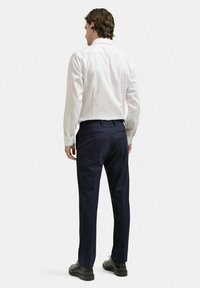 Vit skjorta med långa ärmar, kombinerad med mörkblå byxor. Modellen visas bakifrån, bärandes svarta skor.