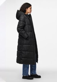 Casaco longo acolchoado preto com capuz, apresentando um design acolchoado, fecho de zip e bolsos laterais. O modelo usa jeans azuis e botas pretas.