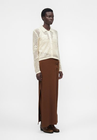 Γυναίκα που φοράει μια κρεμ ανοιχτή πλεκτή ζακέτα με κουμπιά πάνω από μια καφέ μακριά φούστα με ψηλή μέση και ανοίγματα στο πλάι και καφέ μποτάκια μέχρι τον αστράγαλο.