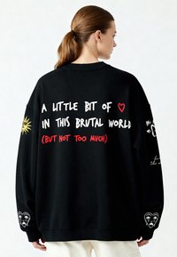 Černá oversized mikina s bílým a červeným textem na zádech, sluneční grafikou na jednom rukávu a designem srdce se slzami na druhém rukávu.