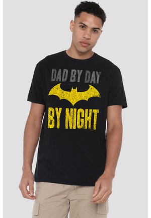 Junger Mann trägt ein schwarzes T-Shirt mit gelbem Fledermaus-Symbol und dem Text "Dad by day, by night" im distressed Stil.